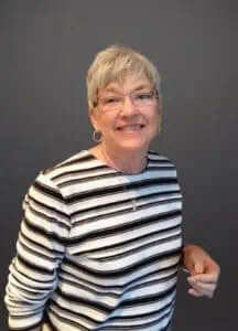 Senior woman with short gray hair and glasses, wearing a striped sweater, smiling warmly against a neutral gray backdrop; representation of compassionate funeral service offerings like cremations and pre-planning at Alabama Funeral Homes.