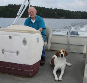 An elderly man navigating a boat on a serene lake, accompanied by a resting brown and white dog; lush trees create a peaceful backdrop. Scene embodies tranquility and reflection for funeral planning and services in Alabama.