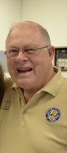 A smiling middle-aged man with glasses in a beige polo shirt featuring a circular embroidered logo, symbolizing professionalism and warmth, ideal for funeral home services in Alabama specializing in cremations, funerals, and pre-planning.